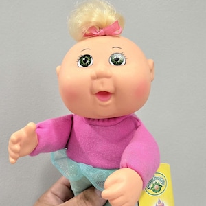 May include: A Cabbage Patch Kids doll with a fair complexion, green eyes, and a small blonde bun with a pink bow. The doll wears a pink long-sleeved shirt and a blue tulle skirt. A yellow tag is visible.