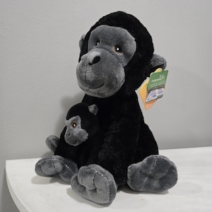 以下が含まれることがあります： ぬいぐるみゴリラと赤ちゃんゴリラのぬいぐるみ。大人のゴリラは黒色で、灰色の顔の特徴と手足があります。赤ちゃんゴリラは黒色で、灰色の顔の特徴があります。どちらのおもちゃも白い面に座っています。