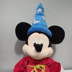 May include: A plush Mickey Mouse dressed as a sorcerer. He is wearing a blue hat with stars and a crescent moon, a red robe, and a gold rope belt.