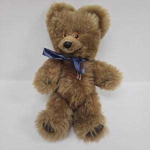 May include: A brown teddy bear with orange eyes and a blue ribbon around its neck. The bear is sitting on a white background.