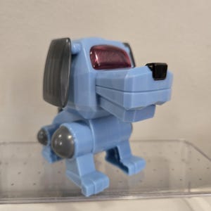 以下が含まれることがあります： 幾何学的でブロック状のデザインをした、水色のプラスチック製犬のフィギュア。このおもちゃには、ダークグレーの耳、脚のダークグレーの関節、小さな黒い鼻、そして目の部分を覆う半透明の赤紫色のバイザーが特徴として備わっています。