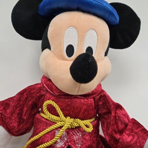 Pu&ograve; includere: Un Mickey Mouse di peluche che indossa una veste rossa con bordi dorati e un cappello blu. La veste ha un motivo paisley e il cappello &egrave; di raso.