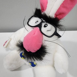 May include: White plush bunny with black glasses, a black moustache, and a pink nose. The bunny is wearing a blue and yellow bow tie.
