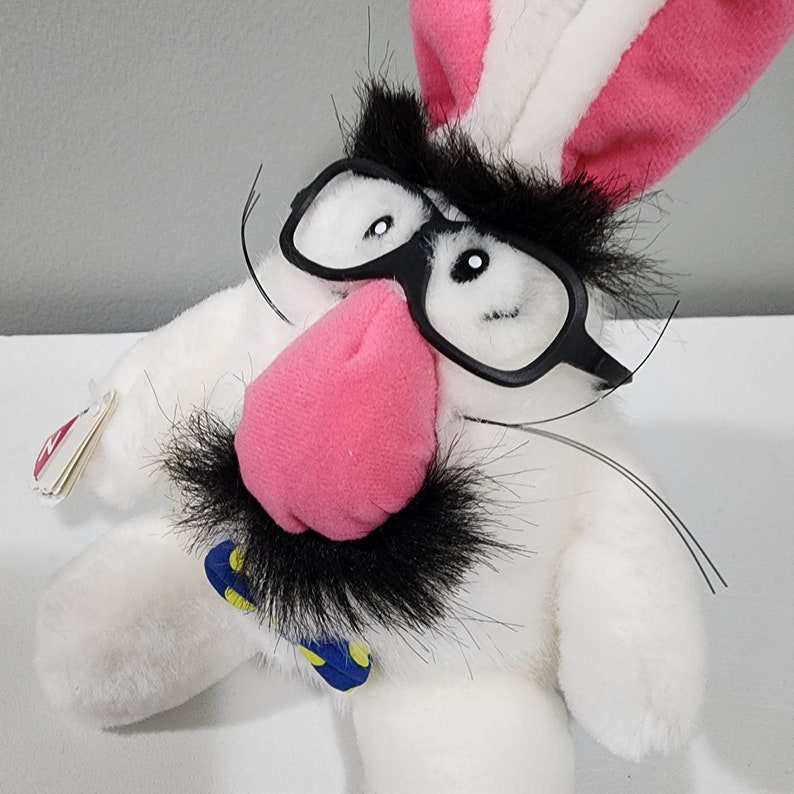May include: White plush bunny with black glasses, a black moustache, and a pink nose. The bunny is wearing a blue and yellow bow tie.
