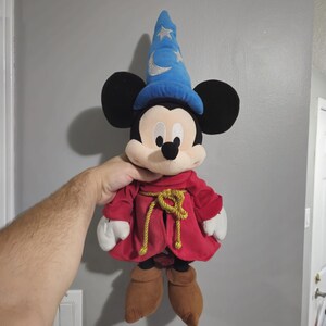 May include: A plush Mickey Mouse toy wearing a blue wizard hat with stars and a red robe with a gold rope belt.