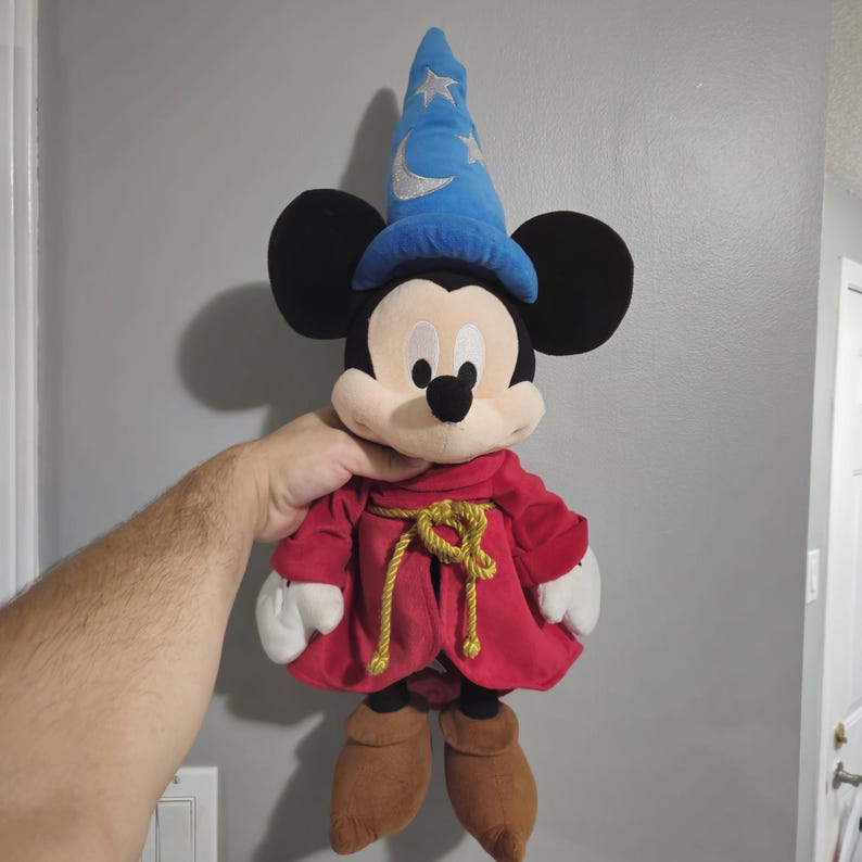 May include: A plush Mickey Mouse toy wearing a blue wizard hat with stars and a red robe with a gold rope belt.