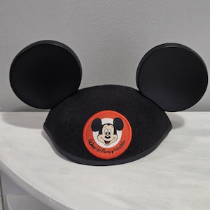 Puede incluir: Sombrero negro con orejas de Mickey Mouse. Cuenta con un parche circular con Mickey Mouse y las palabras "Walt Disney World" en un círculo rojo y blanco. El sombrero tiene dos grandes orejas negras redondas.