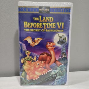 May include: A VHS tape for the animated film "The Land Before Time VI: The Secret of Saurus Rock". The cover features a purple sky with a full moon, a green dinosaur, a brown dinosaur, and a pterodactyl.