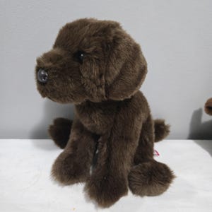 May include: A soft, brown Labrador puppy dog toy. The toy has a black nose and is sitting upright. The puppy's fur is a rich, chocolate brown colour, and it has a friendly, inviting appearance. The toy is made of a soft, furry material.