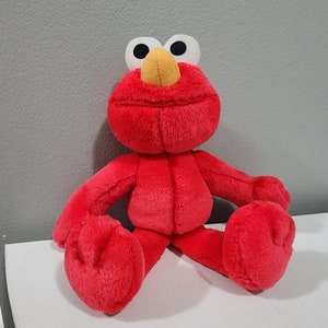 以下が含まれることがあります： 大きな笑顔、大きな白い目、明るいオレンジ色の鼻を持つ赤いぬいぐるみエルモの玩具。おもちゃは白い表面の上に座っています。