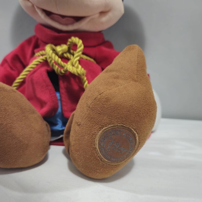 May include: A plush Mickey Mouse toy wearing a red and blue outfit with a gold rope belt. The toy has a brown suede-like finish on its feet and a Disney logo on the bottom of one foot.