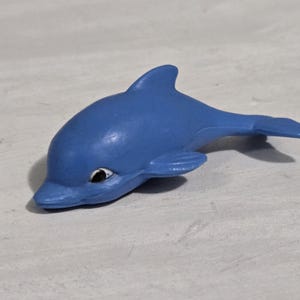 Op de afbeelding: Een blauw dolfijnbeeldje met een wit oog en een gebogen lichaam. De dolfijn ligt op een lichtgrijze ondergrond. Het beeldje is effen van kleur en heeft een gladde textuur.