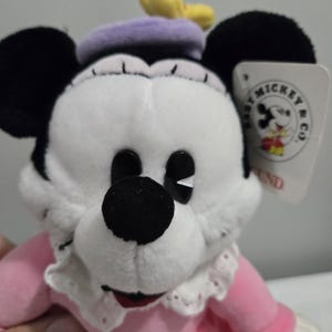 May include: A plush Minnie Mouse doll with a white face, black ears, and a black nose. She wears a pink dress, a purple beret, and a white lace collar. A tag with the words "Baby Mickey & Co" is attached.