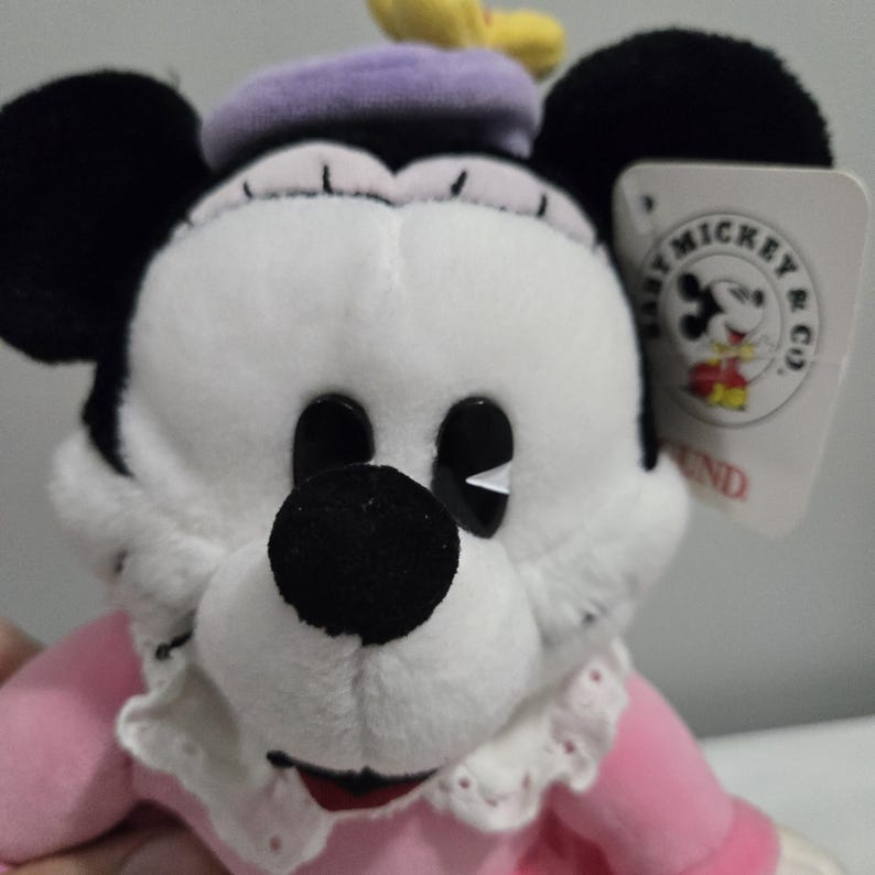 May include: A plush Minnie Mouse doll with a white face, black ears, and a black nose. She wears a pink dress, a purple beret, and a white lace collar. A tag with the words "Baby Mickey & Co" is attached.