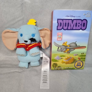 May include: A blue plush Dumbo toy with a red scarf and a yellow hat. The toy is sitting next to a DVD case for the Disney classic movie Dumbo. The DVD case has a picture of Dumbo flying in the air with a circus tent in the background. The text on the DVD case reads "A Walt Disney Classic Dumbo Limited Release - Sortie limitée Dumbo 80".