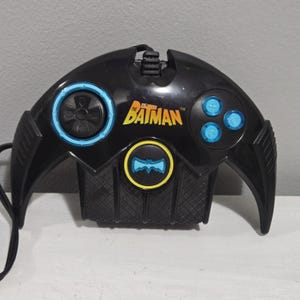 May include: Black Batman video game controller with blue and yellow detailing. The controller has a circular joystick, directional buttons, and the Batman logo. The design is based on the Batman symbol.