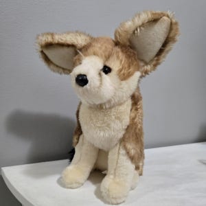 Peut inclure: Un jouet en peluche en forme de chien avec de grandes oreilles dressées. L'animal en peluche a un visage, une poitrine et des pattes de couleur crème, avec de la fourrure marron et beige sur le dos et les oreilles. Il a un nez noir et des yeux sombres.