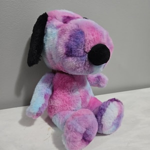 May include: A plush toy dog in a sitting position, featuring a tie-dye pattern in shades of pink, purple, and blue. The dog has black ears and a black nose. The toy is soft and fluffy, with a friendly, cartoonish appearance.