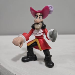 Könnte beinhalten: Eine Captain Hook-Figur. Die Figur trägt einen roten Mantel mit goldenen Verzierungen, eine schwarze Hose und schwarze Stiefel. Er hat einen Haken als Hand und einen rosa Hut mit einer weißen Feder. Die Figur steht in einer dynamischen Pose.
