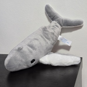 May include: A gray plush whale toy with a white belly and a gray tail fin. The whale has a black eye and a tag that reads "Thelma". The toy is resting on a black surface.