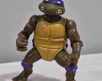 May include: A Donatello action figure from the Teenage Mutant Ninja Turtles franchise. The figure is brown with a yellow plastron and purple accents. It is posed in a fighting stance, with a determined expression.
