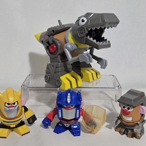 Könnte beinhalten: Eine Sammlung von Spielfiguren, darunter ein grauer und gelber Dinosaurier, ein gelber Roboter, ein blau-roter Roboter und eine braune Figur mit Hut. Die Spielzeuge sind auf einer transparenten Kunststoffoberfläche vor weißem Hintergrund angeordnet.
