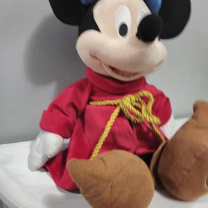 May include: A plush Mickey Mouse wearing a red robe with a gold rope belt. He has a blue hat and brown shoes.