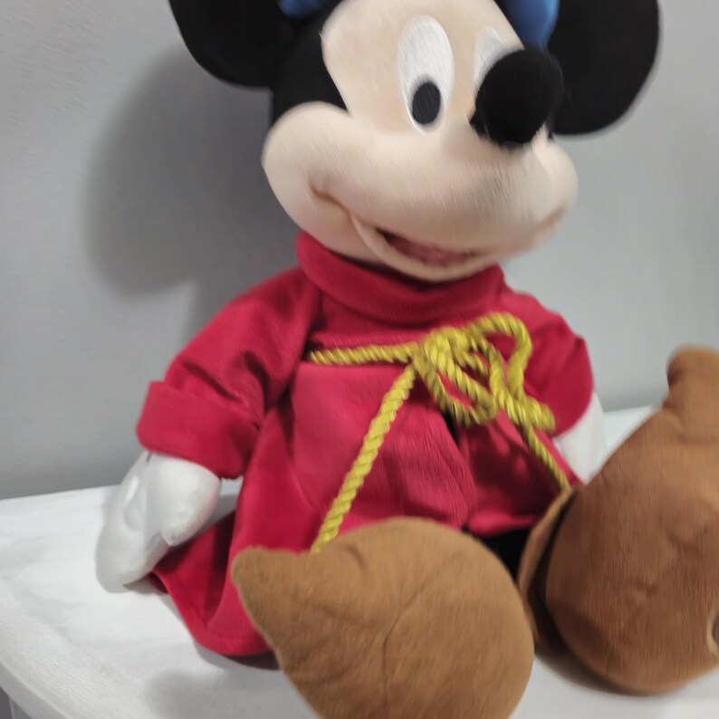 May include: A plush Mickey Mouse wearing a red robe with a gold rope belt. He has a blue hat and brown shoes.