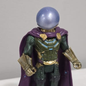 May include: A Mysterio action figure with a blue, spherical helmet and a purple cape. The figure has a green and gold armored suit with blue accents. The arms and gauntlets are gold. The figure is a collectible toy.