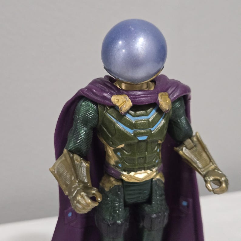May include: A Mysterio action figure with a blue, spherical helmet and a purple cape. The figure has a green and gold armored suit with blue accents. The arms and gauntlets are gold. The figure is a collectible toy.