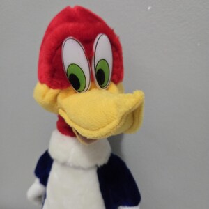 May include: A plush Woody Woodpecker doll. The doll features a red head with a yellow beak, large white eyes with green irises, and a blue and white body. The plush toy is a classic cartoon character.