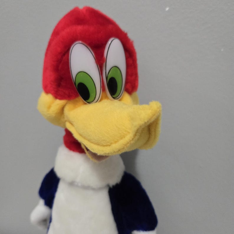 May include: A plush Woody Woodpecker doll. The doll features a red head with a yellow beak, large white eyes with green irises, and a blue and white body. The plush toy is a classic cartoon character.