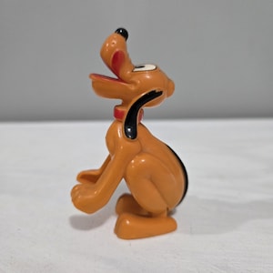 Könnte beinhalten: Eine Vintage-Plastikfigur von Pluto, dem Disney-Hund. Pluto sitzt mit offenem Maul und herausgestreckter Zunge. Er ist braun mit schwarzen Ohren und einem roten Halsband.