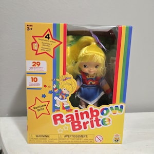 May include: A Rainbow Brite doll in a box. The doll has yellow hair and is wearing a blue dress with a rainbow design. The box is yellow with the words "Rainbow Brite" in colourful letters and includes the text "4 Surprises to unbox!"