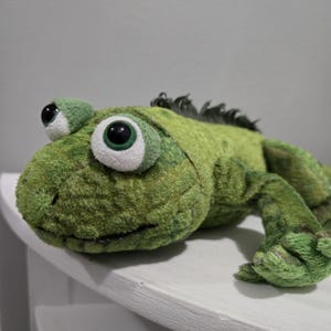 May include: A green plush iguana toy with large eyes and a spiky ridge. The toy is made of a textured, fuzzy material and has a friendly, cartoonish appearance. The iguana is lying down, with its legs extended. A children's toy.