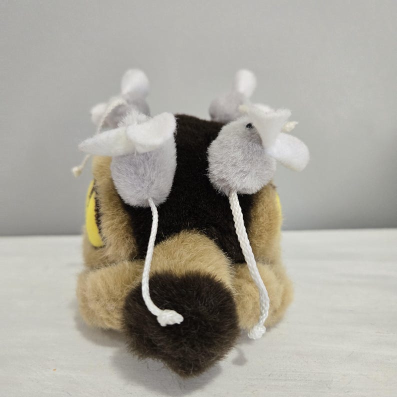 May include: A plush toy cat with a brown and black body, yellow eyes, and white accents. The cat has three gray pom-poms with white petal-like shapes attached by strings. The tail is a dark brown, fluffy shape.