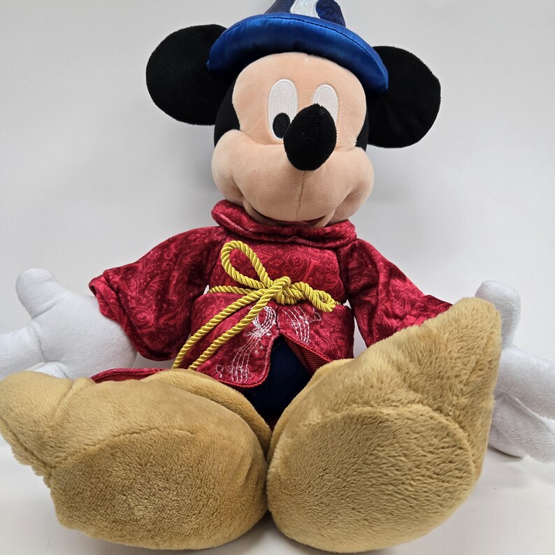 Pu&ograve; includere: Un Mickey Mouse di peluche che indossa una tunica rossa e dorata e un cappello da mago blu.