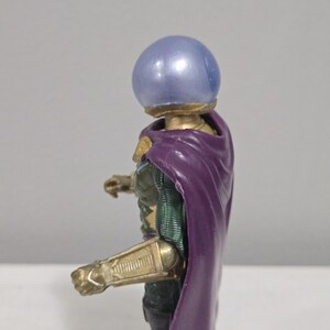 May include: A Mysterio action figure, featuring a metallic gold and green armored suit, a purple cape, and a blue, spherical helmet. The figure is posed in a dynamic stance, showcasing intricate details.