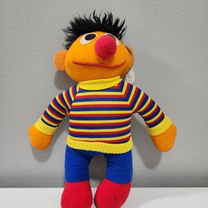 Könnte beinhalten: Ein Plüschtier von Elmo aus der Sesamstraße. Elmo trägt ein blau-gelb gestreiftes Shirt mit gelbem Kragen und blaue Hosen. Elmo hat eine rote Nase und ein schwarzes Augenpflaster.