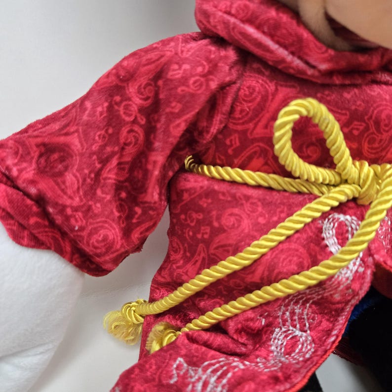 Pu&ograve; includere: Accappatoio di peluche rosso con cintura in corda dorata. L'accappatoio ha un motivo di note musicali bianche.