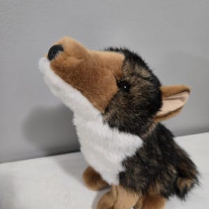 May include: A plush toy shaped like a German Shepherd dog with a tan muzzle, a black nose, and white fur on the chest and chin. The toy features dark brown and black fur on the back and head, with pointed ears and a head tilted upward.