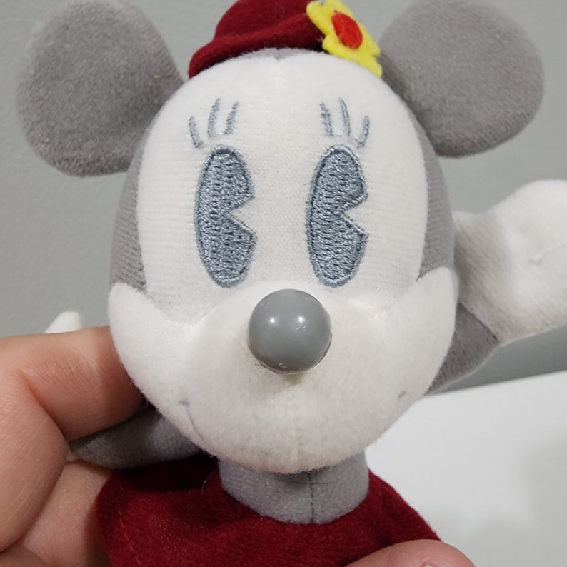 May include: A plush toy mouse with gray ears and a red hat with a yellow flower. The mouse has white fur and gray embroidered eyes. The nose is a gray button.