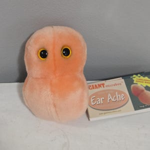 Könnte beinhalten: Ein Plüschtier in Form einer Mikrobe in sanftem Pfirsichton mit zwei großen, runden, schwarzen Augen mit gelben Iris. Neben dem Plüschtier befindet sich ein rechteckiges Produktetikett mit dem Text GIANT microbes und Ear Ache auf der Vorderseite.