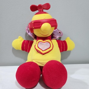May include: A plush toy character with a yellow body, red accents, and red sunglasses. The toy has a heart-shaped emblem on its chest and large red feet. It has a red propeller on top of its head and a purple collar.
