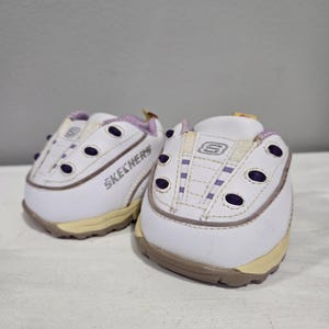 May include: A pair of white Skechers slip-on shoes with purple accents and a yellow tag. The shoes have a white leather upper with a textured design and a white rubber sole.