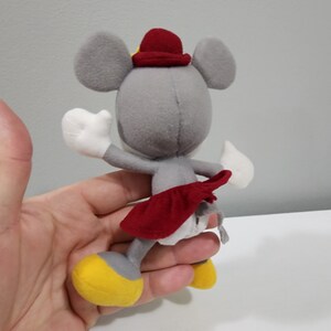 May include: A gray plush mouse wearing a red hat and a red skirt. The mouse is standing on its hind legs and has its arms outstretched.