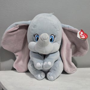 May include: A plush Dumbo the Elephant toy in a seated position. The toy is primarily light blue with large, pink-lined ears. It has blue eyes, a gray trunk, and white feet. A red Ty tag is attached to the ear.