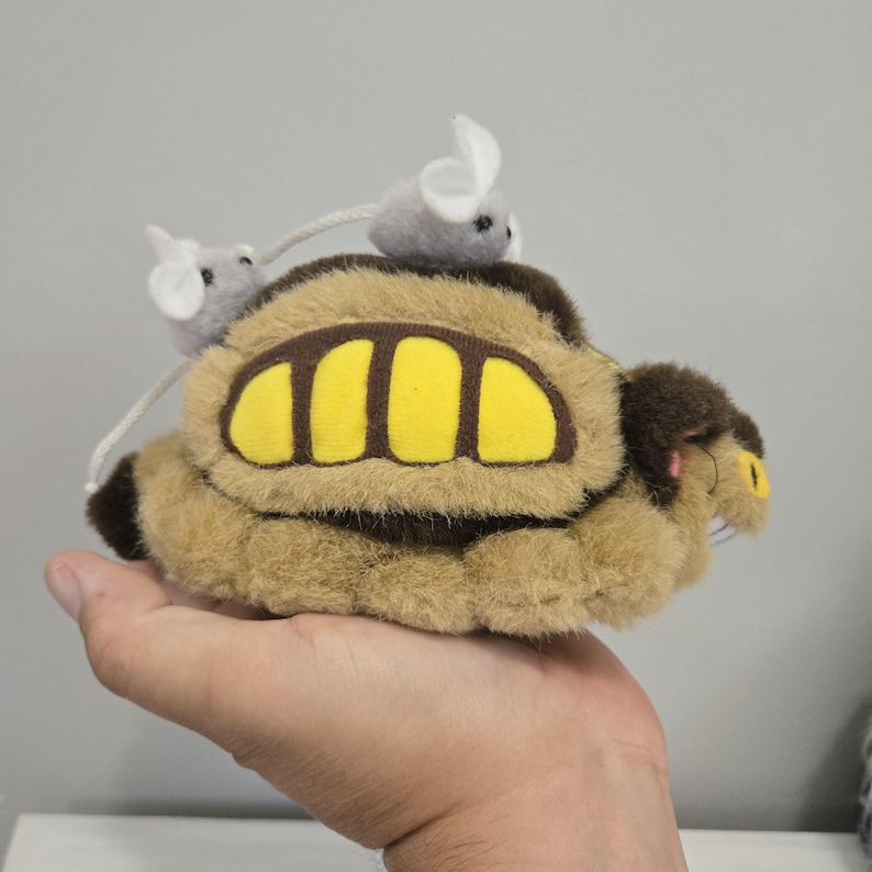 May include: A plush toy resembling a character from an animated film, featuring a brown body with yellow accents and two small gray mice on top. The toy is held in a hand.