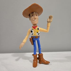 Pu&ograve; includere: Una figurina di plastica di Woody del franchise Toy Story. Woody indossa il suo cappello da cowboy marrone distintivo, un gilet giallo con una bandana rossa, jeans blu e stivali marroni. Sta salutando con la mano destra.