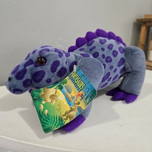 May include: A purple and gray plush dinosaur with a tag that reads "Fantasaurus Collection 24/C the Company".